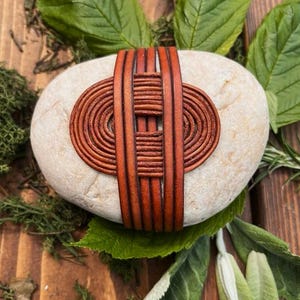 May include: An oval, pale stone with a brown leather design. The leather is woven in a circular and linear pattern. The stone is surrounded by green leaves and moss, set on a wooden surface.