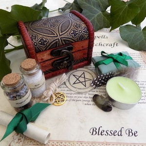 May include: A small wooden chest with a gold and black floral design, two small glass bottles with cork tops, a green ribbon, a rolled up piece of parchment, a small white candle, a green and brown feather, a small green stone, a gold pentagram charm, and a piece of twine. The text "Spells & Witchcraft" and "Blessed Be" are visible on the background.