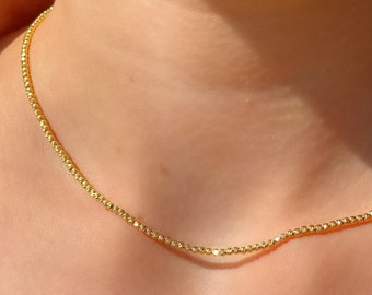 May include: A delicate gold necklace with a beaded design. The necklace is worn around the neck, and the gold color reflects the light. The necklace is a simple yet elegant piece of jewelry.