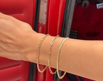 May include: Three gold chain bracelets on a person's wrist. The bracelets are different styles, including a curb chain, a rope chain, and a paperclip chain.