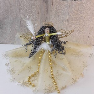 Op de afbeelding: Handgemaakte engelenfiguur met een gouden kroon, grijs haar en een gouden en witte jurk. De engel heeft gouden vleugels en is versierd met gouden kralen en witte veren. De tekst "Jean's crafts" is zichtbaar.
