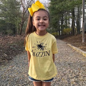 Puede incluir: Una niña pequeña con una camiseta amarilla con un gráfico de abeja negra y el texto "Buzzin". Lleva un lazo amarillo en el pelo y está de pie en un camino de grava.