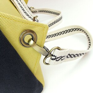 May include: Close-up of a yellow and black canvas tote bag with a brass buckle and a white strap with black stitching.