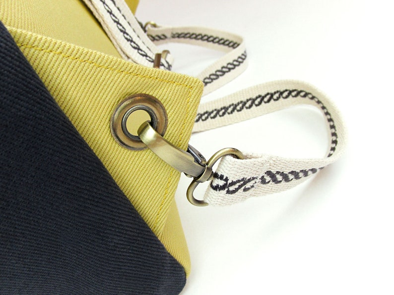 May include: Close-up of a yellow and black canvas tote bag with a brass buckle and a white strap with black stitching.