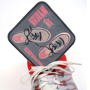 May include: A grey and pink embroidered patch with the text "Walk It Out" and a pair of pink trainers with black laces.