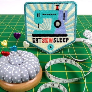 Może przedstawiać: Kolorowa łatka z maszyną do szycia z tekstem "Eat Sew Sleep" na zielonej macie do cięcia z poduszką na szpilki i miarą krawiecką.