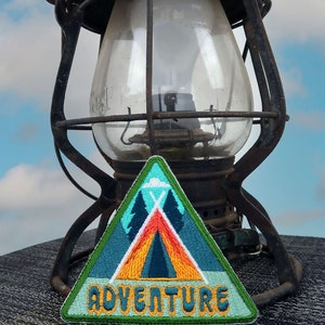 Könnte beinhalten: Ein grüner, blauer und orangefarbener gestickter Patch mit einem Zelt und Bäumen. Der Patch ist dreieckig und hat das Wort "ADVENTURE" in gelber Schrift.