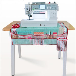 May include: A teal and red sewing machine mat and caddy with pockets for storing sewing supplies. The mat is on a wooden table with a white sewing machine on top.