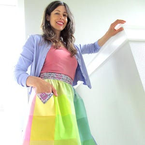 May include: A woman wearing a light blue cardigan over a pink tank top and a colorful patchwork skirt with a small pocket. The skirt is made of squares of different shades of pink, yellow, and green.