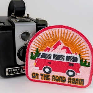 May include: A pink and orange embroidered patch with a red camper van, mountains, and a sun. The text "On the Road Again" is written in black and orange letters.