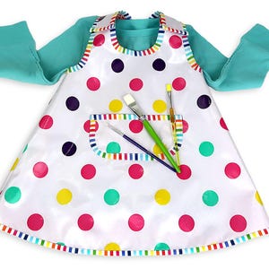 May include: A white art smock with colorful polka dots and a rainbow-striped trim. The smock has a pocket and is paired with a teal long-sleeved shirt. Paintbrushes are in the pocket.