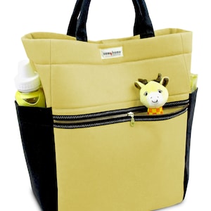 May include: A beige canvas tote bag with black trim and a zippered pocket. The bag has a black handle and a small yellow and orange stuffed animal attached to the zipper. The bag has the text "sew4home" printed on the front.