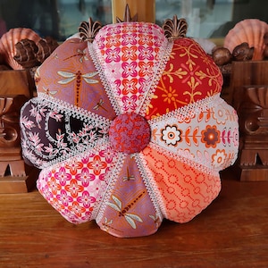 May include: A flower-shaped cushion made from segments of patterned fabric. The cushion has a central button and is edged with white lace. The fabrics feature floral and dragonfly designs in pink, orange, red, and brown hues. The cushion is a decorative item.