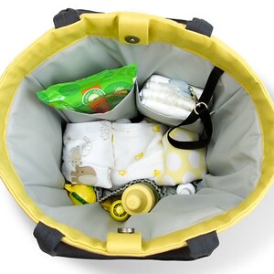 May include: A yellow and black canvas tote bag with a gray lining. The bag is filled with baby items, including diapers, wipes, a bottle, and a few outfits.