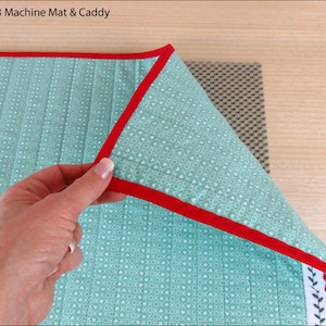 May include: A turquoise blue machine quilting mat with a red binding and a white and green heart pattern. The mat is folded over a gray non-slip mat.