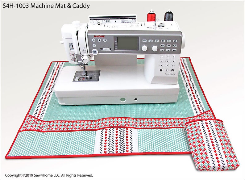 May include: A white sewing machine with a teal and red patterned mat and a matching caddy. The mat has pockets for storing sewing supplies. The machine has a digital display and a variety of buttons. The text "JANOME" is visible on the machine.