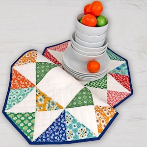 Könnte beinhalten: Ein bunter Patchwork-Tischläufer mit einem marineblauen Rand. Der Läufer besteht aus verschiedenen Stoffen mit floralen und geometrischen Mustern. Ein Stapel weißer Teller und Schalen mit Orangen darauf befinden sich auf dem Läufer.