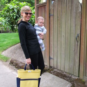 May include: A woman wearing sunglasses and a black long-sleeved shirt is holding a baby wearing a striped onesie. The woman is carrying a yellow and blue tote bag with a zipper.