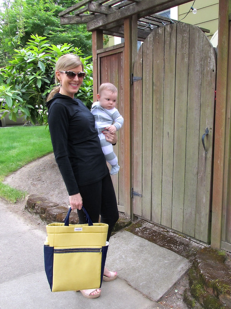 May include: A woman wearing sunglasses and a black long-sleeved shirt is holding a baby wearing a striped onesie. The woman is carrying a yellow and blue tote bag with a zipper.