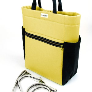 May include: A yellow tote bag with black trim and a black zippered pocket. The bag has a black handle and a detachable shoulder strap with a white and black braided design.