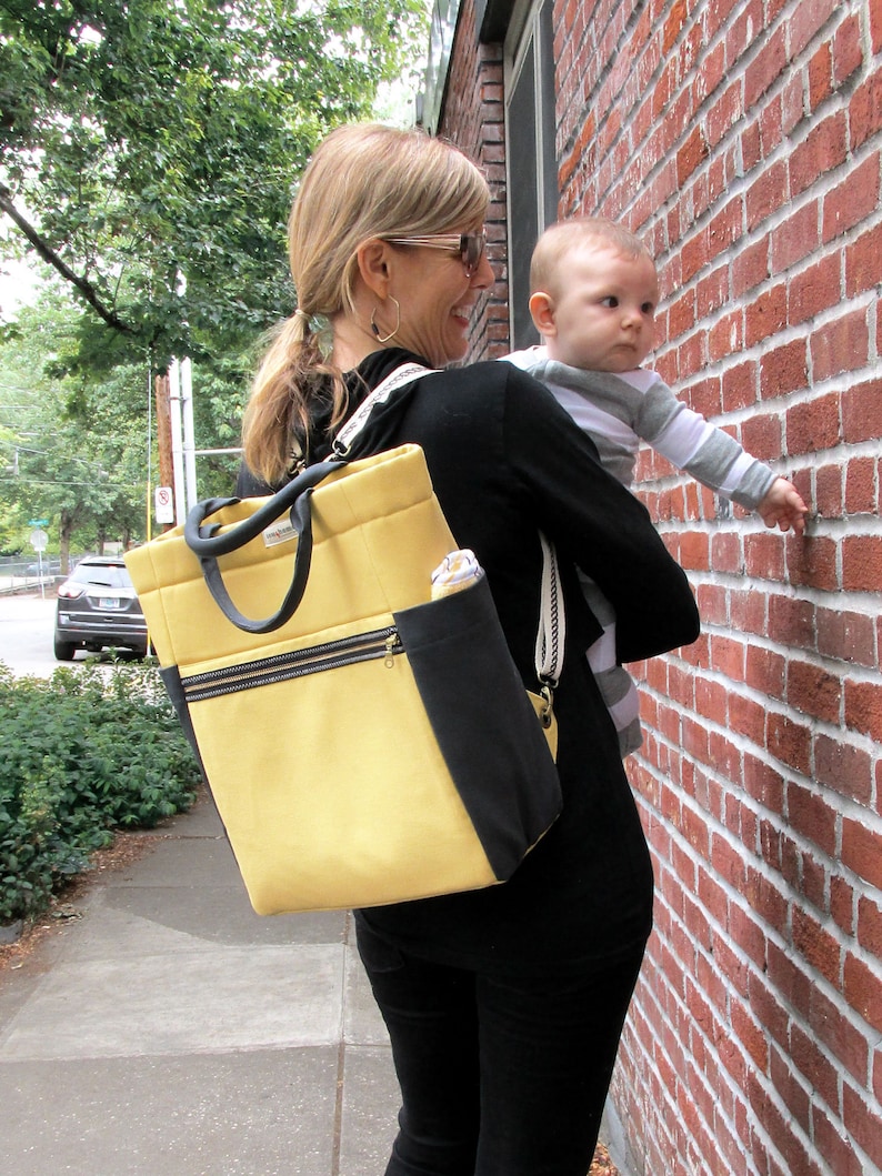 May include: A woman wearing a black shirt and a yellow and black backpack is carrying a baby. The baby is wearing a white and gray striped shirt and is looking at the camera.