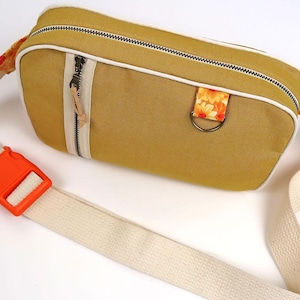 May include: Mustard-colored fanny pack with a white trim and a cream-colored strap. The bag features a black zipper, a small leather pull, and an orange buckle. A floral fabric detail is attached to the bag.