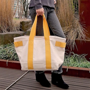 May include: Cream-colored tote bag with bright yellow straps and accents. The bag is rectangular with a flat bottom and is being held by a person wearing dark jeans and black boots. The bag has a simple, clean design.