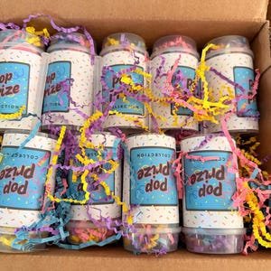 May include: A cardboard box filled with cylindrical containers labeled "Pop Prize Collection." The containers are white with a blue label and colorful confetti design. Yellow, purple, and pink paper streamers surround the containers.