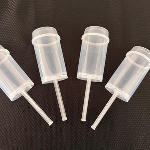 May include: Four clear plastic push pop containers with white stems and a white ring around the top. The containers are cylindrical and are arranged on a dark surface.