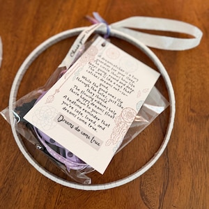 May include: A white dreamcatcher hoop with a clear plastic bag containing purple ribbons and a card with text. The card reads "Dreams do come true" and includes a poem about dreams. The dreamcatcher is on a wooden surface.
