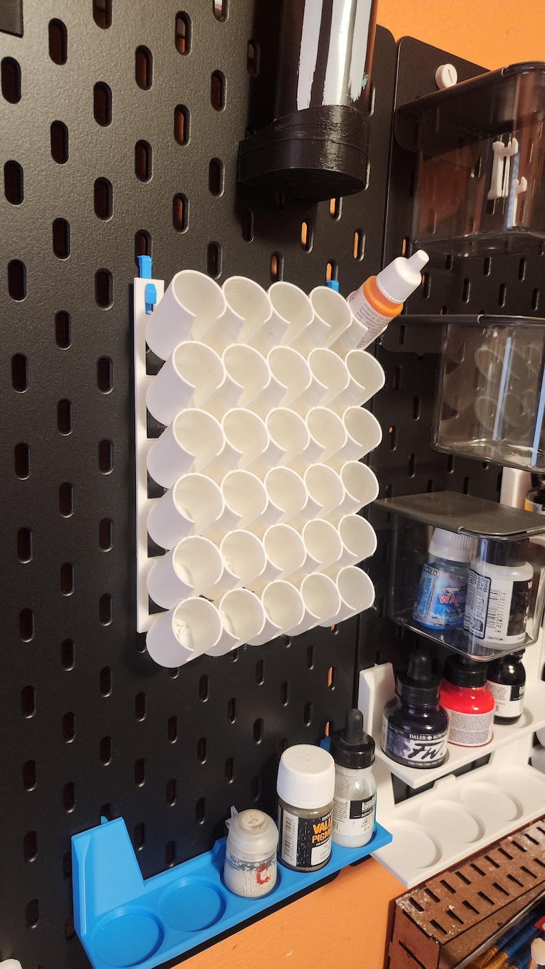 May include: A white plastic paint rack with 24 round holes for holding paint bottles. The rack is mounted on a black pegboard wall. There are several bottles of paint and other painting supplies on shelves below the rack.