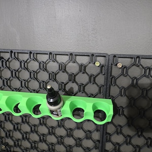 May include: A black, geometric wall-mounted organizer with a bright green paint bottle holder. The holder is designed to hold several small paint bottles. A single paint bottle is in the holder. The organizer is attached to the wall with screws.