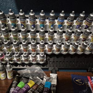 May include: A selection of miniature paint bottles in a white and black organiser. The bottles are arranged in rows and columns, with various colours visible. Tubes of acrylic paint and a logo are also present.