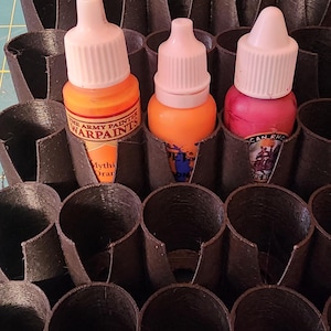 May include: A black plastic paint pot holder with three bottles of paint. The bottles are labelled "The Army Painter Warpaints" and have orange and pink paint.