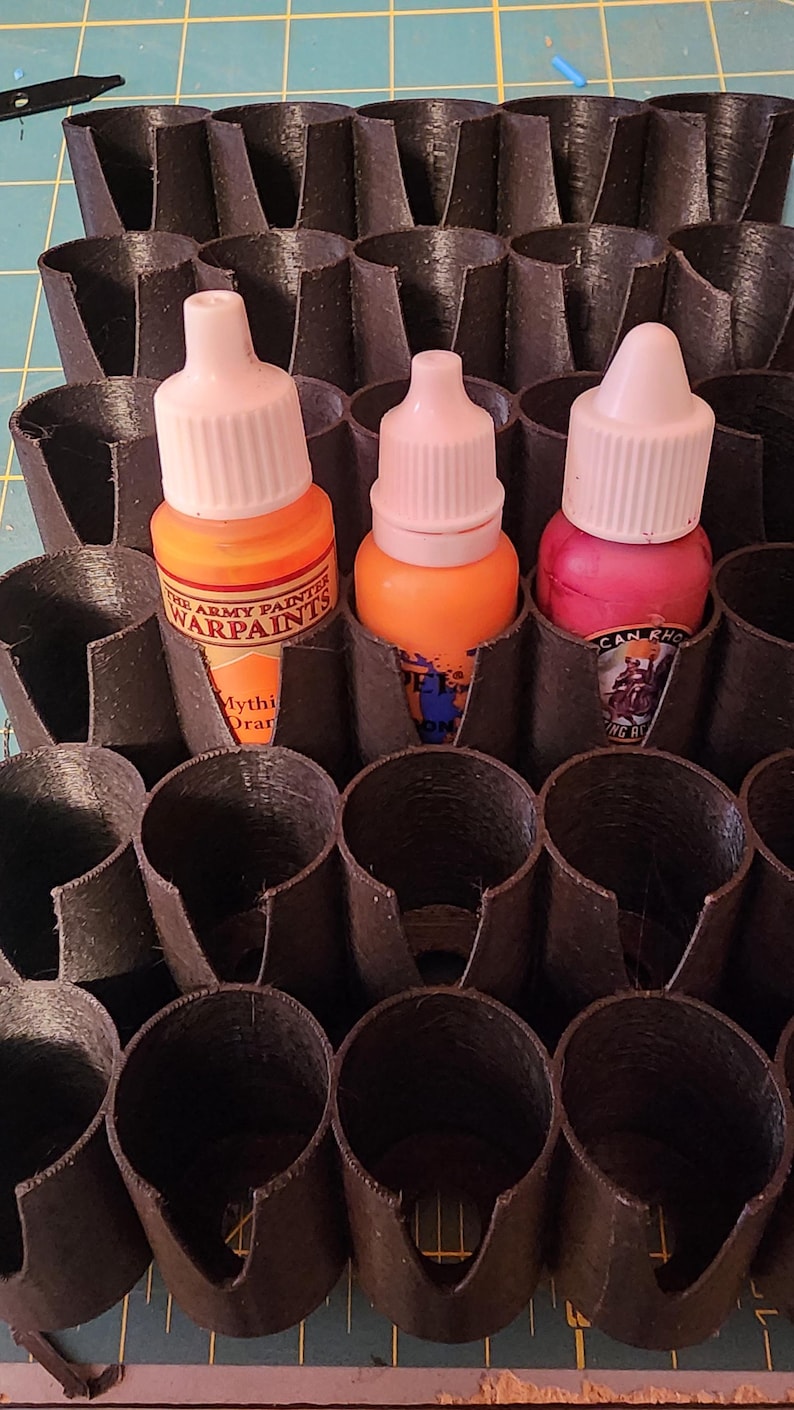 May include: A black plastic paint pot holder with three bottles of paint. The bottles are labelled "The Army Painter Warpaints" and have orange and pink paint.