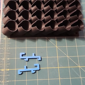 May include: A black 3D printed object with a honeycomb pattern. The object is made up of many small, interconnected cylinders. There are two blue plastic pieces on a green cutting mat.