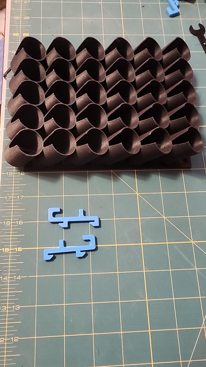 May include: A black 3D printed object with a honeycomb pattern. The object is made up of many small, interconnected cylinders. There are two blue plastic pieces on a green cutting mat.