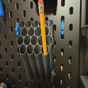 Puede incluir: Un porta lápices negro impreso en 3D con 18 ranuras para lápices. Un lápiz amarillo está en una de las ranuras.
