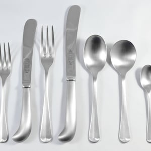 Puede incluir: Un juego de cubiertos de plata, que incluye un cuchillo para mantequilla, un tenedor, una cucharita y dos cucharas de sopa. Los cubiertos están dispuestos en una fila sobre una superficie blanca.