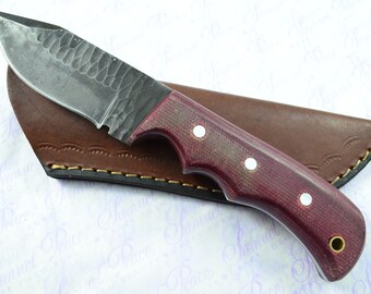 May include: A handmade knife with a dark metal blade and a burgundy handle with white accents. The knife is in a brown leather sheath.