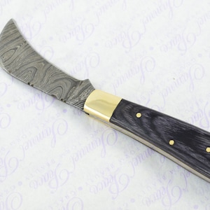 May include: A black handled folding knife with a Damascus steel blade. The blade has a curved edge and a small serrated section. The handle has brass accents.