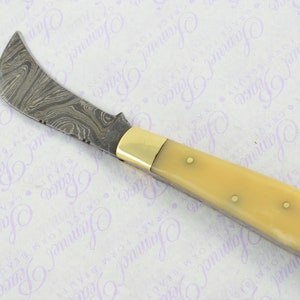 May include: A curved, stainless steel knife with a patterned blade and a cream-colored handle. The handle has a brass accent and a small hole at the end.