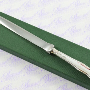 May include: A silver letter opener with a long, thin blade and a curved handle. The handle is engraved with the words "Gorham Sterling". The letter opener is resting on a green box.