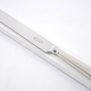 Può includere: Un coltello da torta d'argento con una lama lunga e sottile e un manico arrotondato. Il coltello ha l'iscrizione "The Queen" sulla lama.