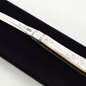 May include: A silver and mother-of-pearl pocket knife with a decorative handle and a hook-shaped blade. The knife is marked with the letters "OW&P" and the number "033".