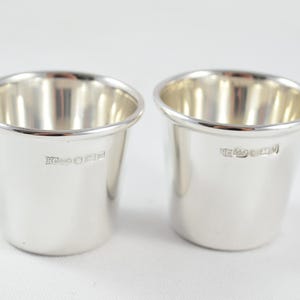 Può includere: Una coppia di bicchieri in argento lucido con una forma cilindrica affusolata e liscia e un bordo arrotondato. Ogni bicchiere presenta un piccolo marchio stampato vicino al bordo superiore, che indica la qualità del metallo e l'origine.