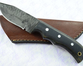 May include: A black handled knife with a hammered steel blade and a brown leather sheath. The handle has white dots.