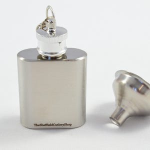 Puede incluir: Un petaca de acero inoxidable con tapón de rosca y llavero. Un pequeño embudo está al lado de la petaca. La petaca es rectangular y tiene el texto "TheSheffieldCutleryShop" impreso en la parte inferior.