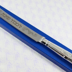 May include: A silver bread knife with an ornate handle and a serrated blade. The blade is engraved with the Hebrew words "L'Chaim" which means "To Life!" The knife is in a blue box.
