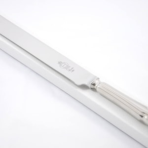 Può includere: Un coltello da torta argentato con una lama lunga e piatta e un manico decorativo. Il coltello ha una superficie lucida e riflettente ed è posto su uno sfondo bianco. Il manico ha un design a coste e un'estremità curva.